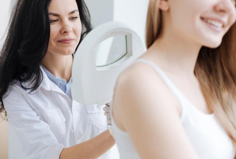 A dermatologist holding a handheld mirror for a young woman, who smiles as she examines her reflection, in a well-lit clinical environment.