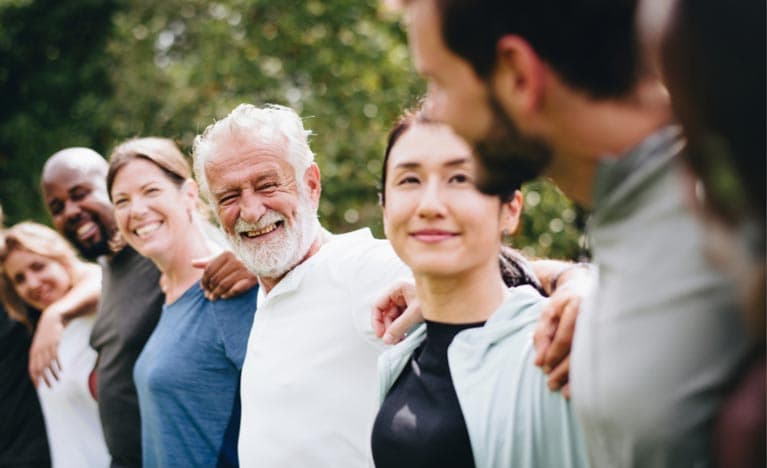 A diverse group of happy people standing arm-in-arm in a park, including an elderly white man with a white beard smiling joyfully, flanked by a Black man, a white woman, and an Asian woman, with a focus on community and togetherness.