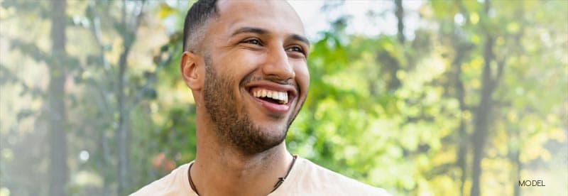 Close-up of a joyful young man with a shaved head, smiling broadly in a sunlit park, surrounded by lush greenery.